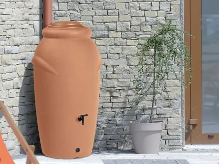 Nádoba na sběr dešťové vody u kamenné zdi s rostlinou.